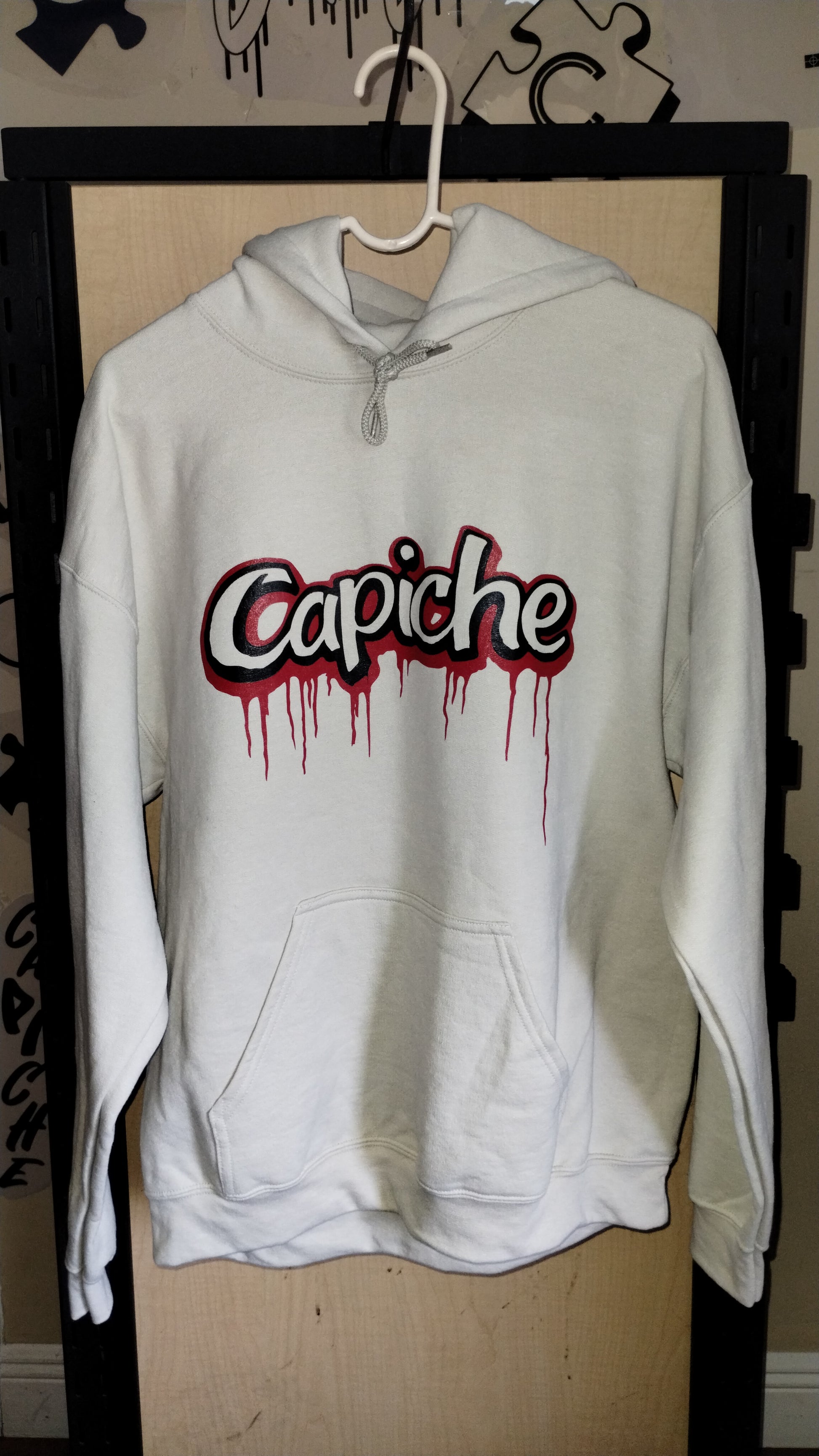 Front view of tan hoodie on a hanger. Two tone Capiche logo in black and red ink.