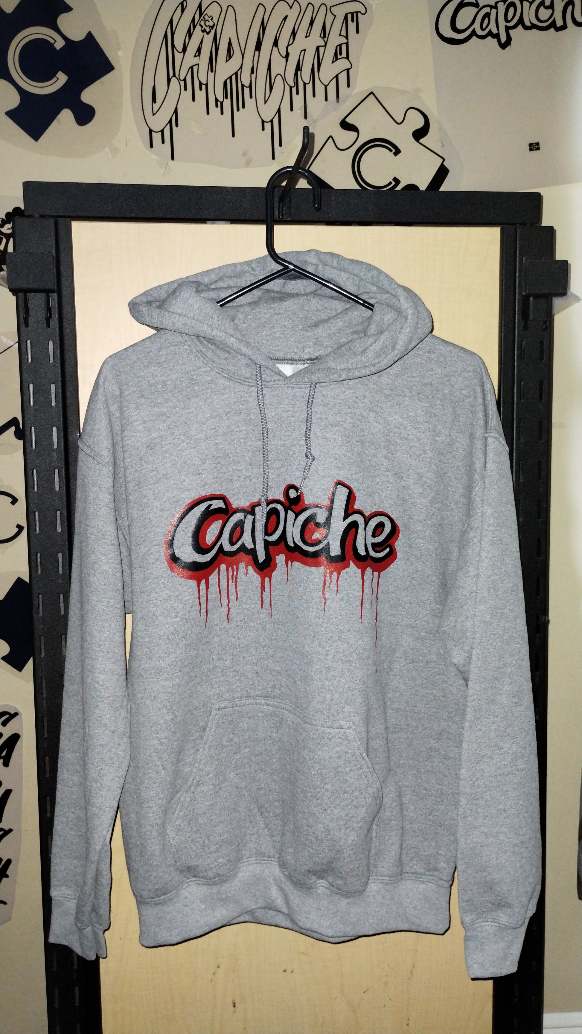 Front view of grey hoodie on a hanger. Capiche logo in black and red ink.