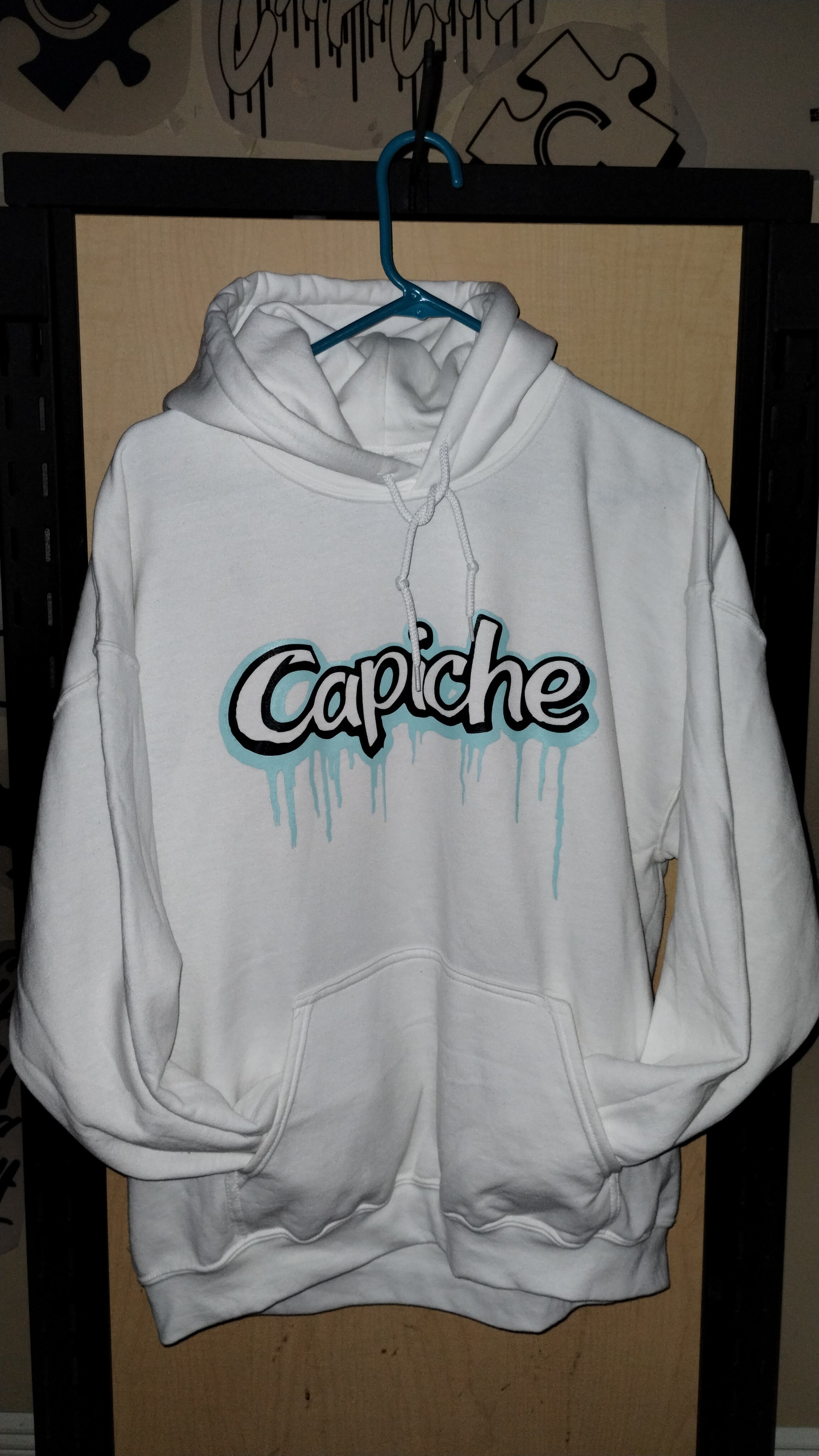 Front view of a white hoodie on a hanger. Capiche logo in black and teal ink.