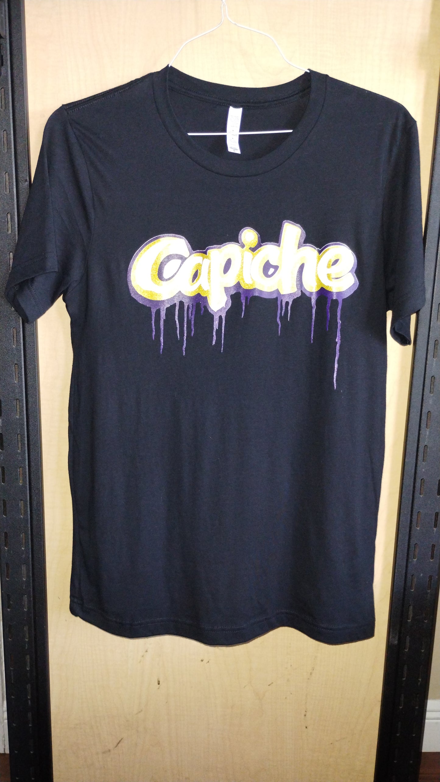 Front view of black t-shirt on a hanger. Capiche printed in white, gold, and purple inks.