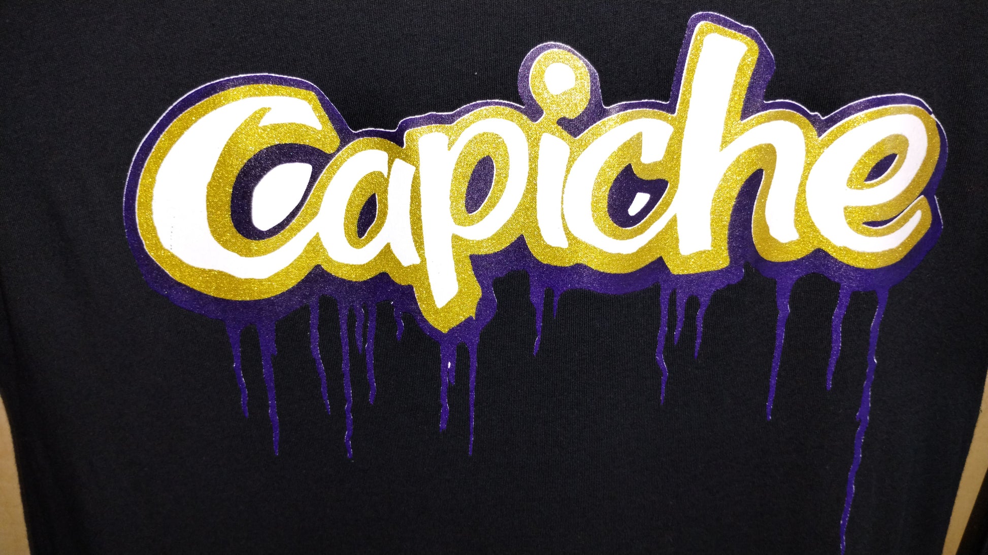 Focused shot of Capiche logo on black shirt. Logo is in white, gold and purple inks.