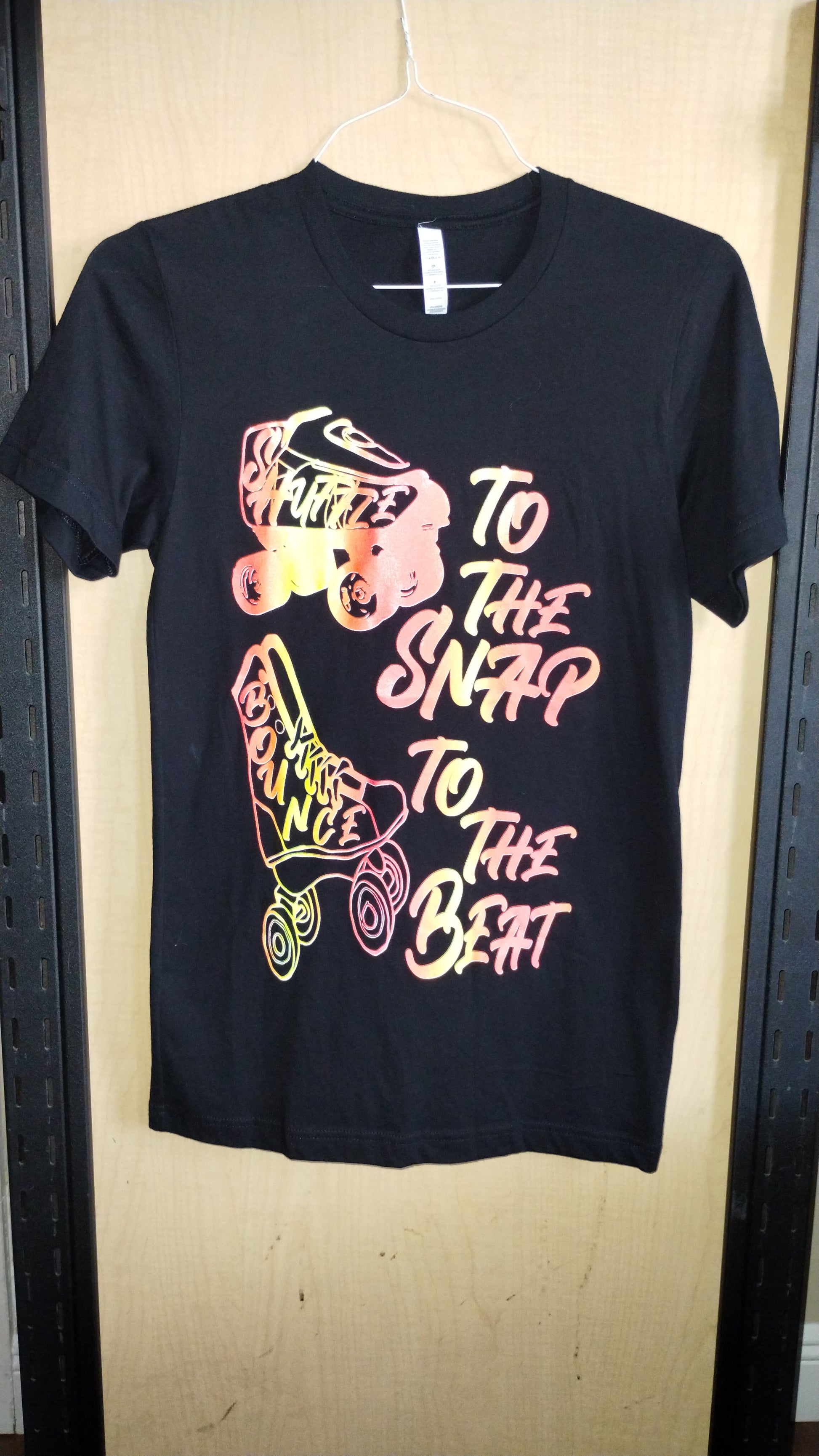 Front view of black shirt on a hanger. Drawing of two different kind of skates with text stating Shuffle to the Snap Bounce to the Beat.